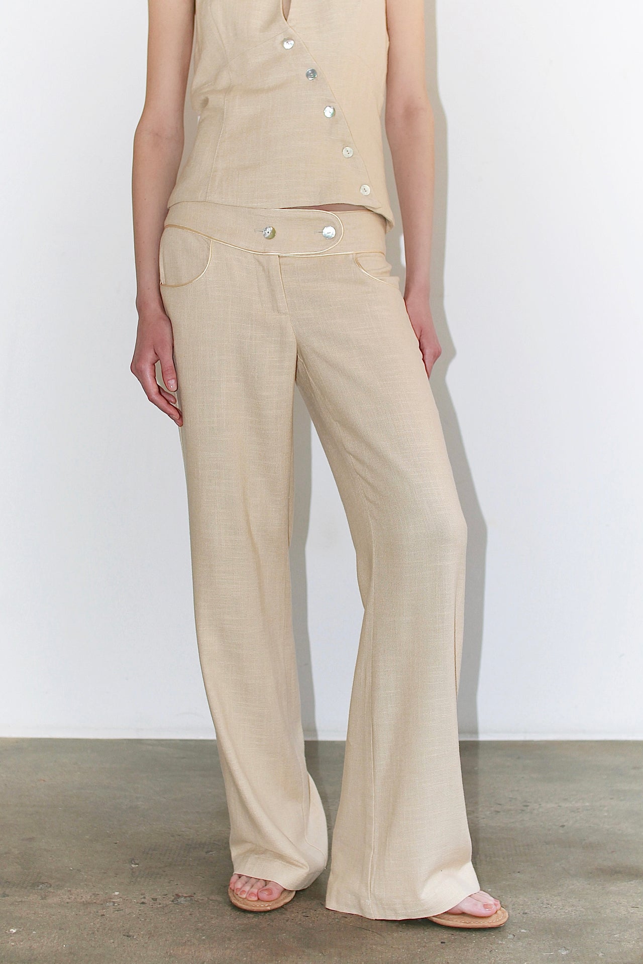 Front view of the Andrea Pants in warm beige Italian linen with contrasting piping, featuring a low-rise fit, curved waistband, and mother-of-pearl button detail.
