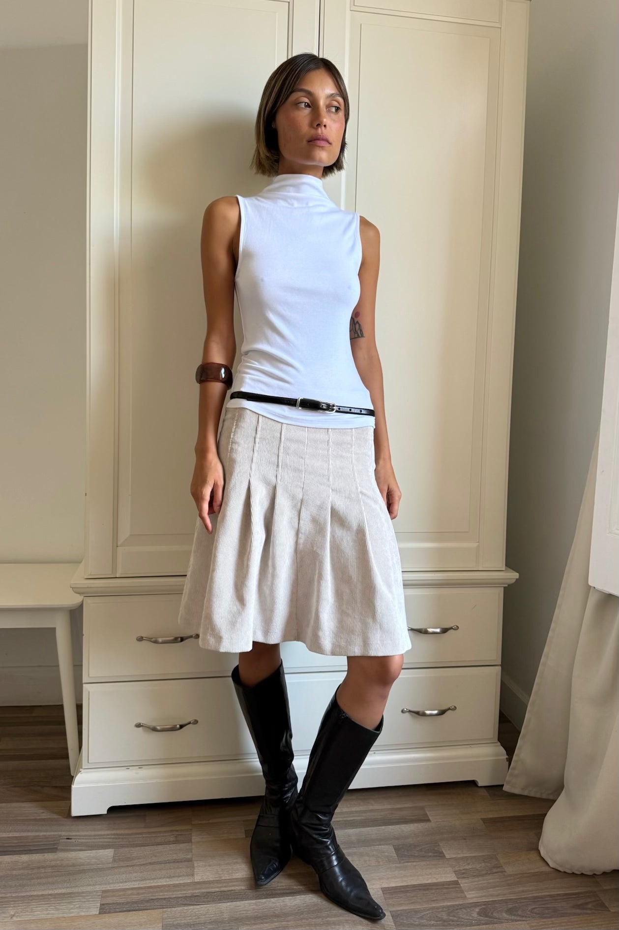 Evie ivory pleated skirt worn with a white top and black boots indoors